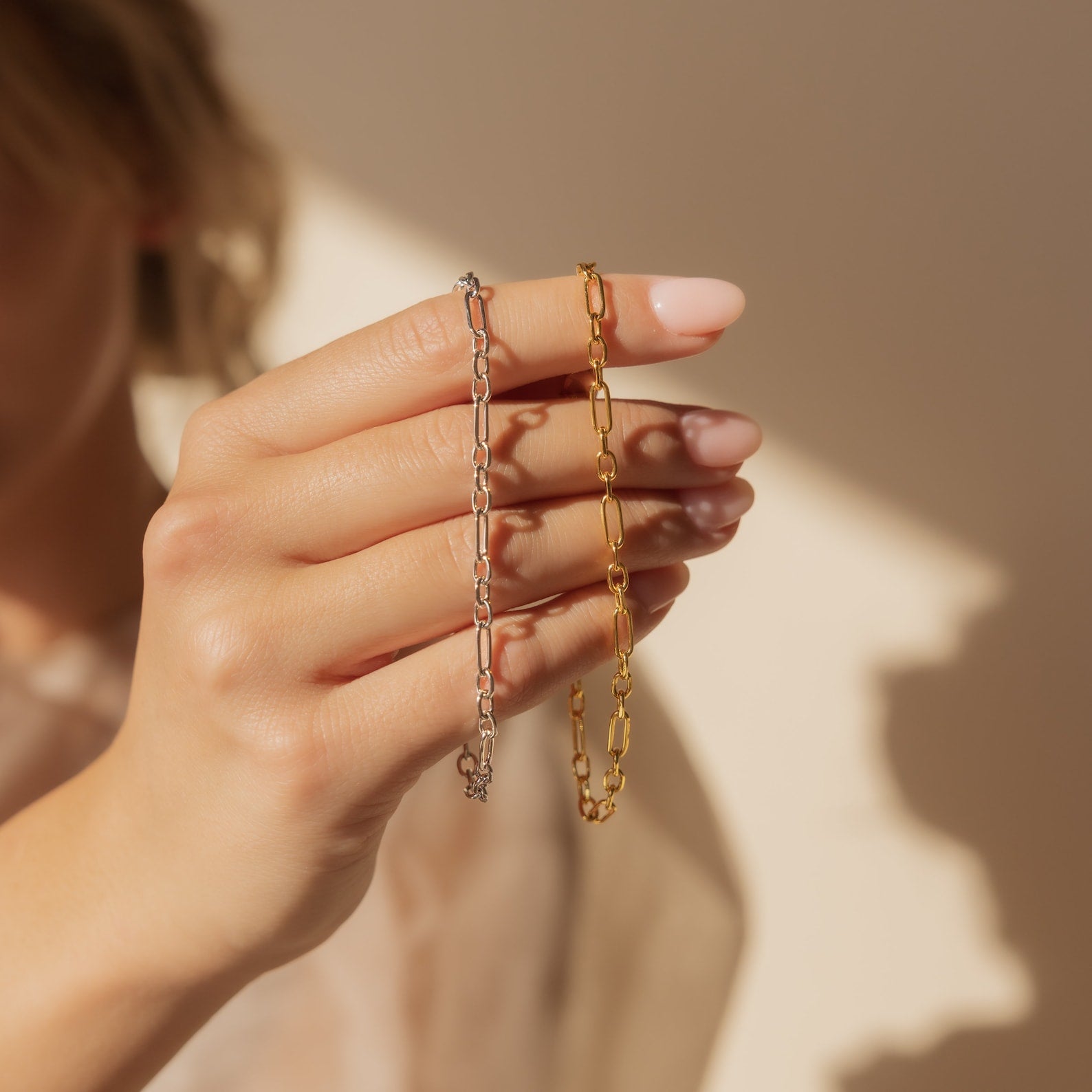 A hand displays a minimal jewelry collection featuring the Roxy Paperclip Chain Bracelet and a gold chunky bracelet against a soft, neutral background.