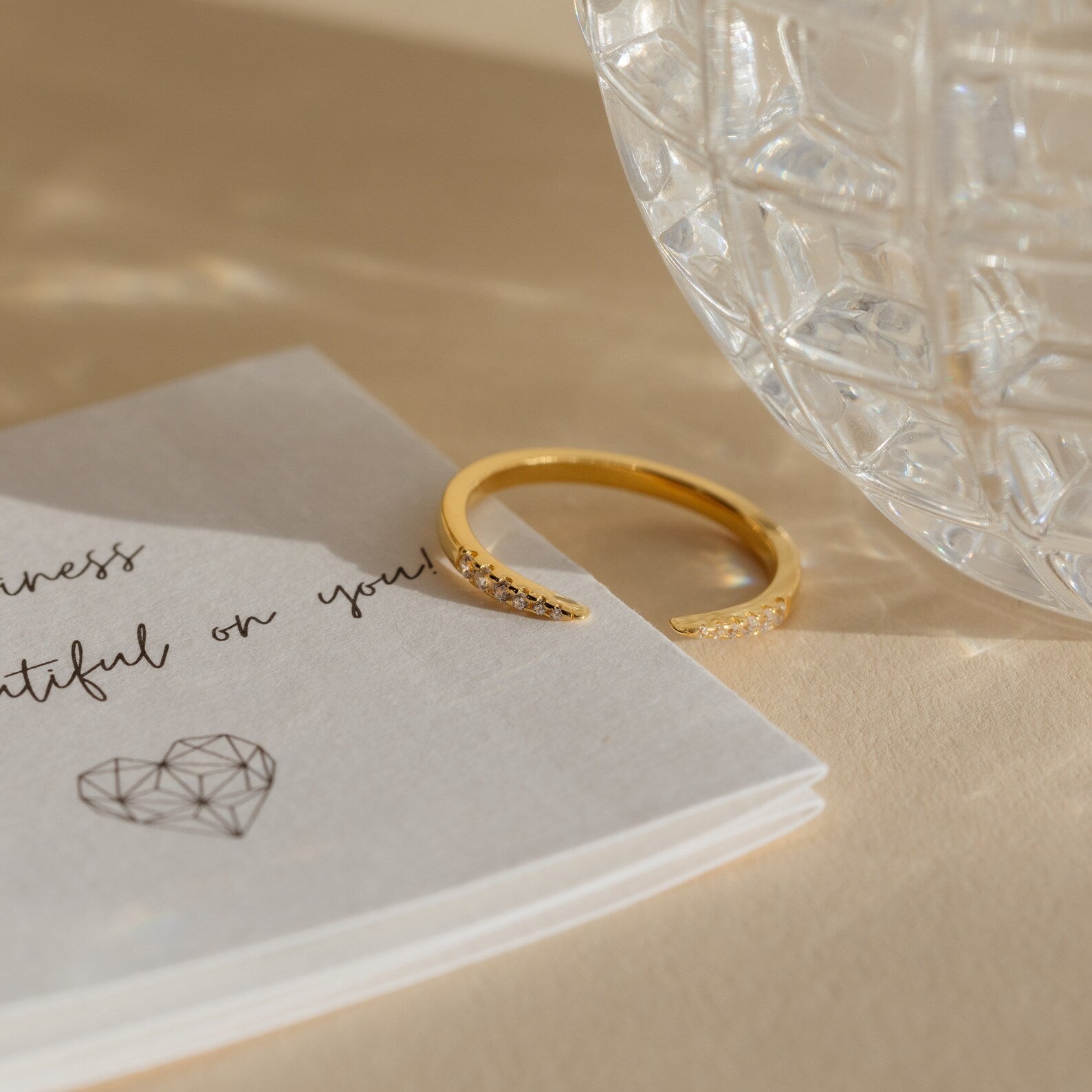 The Jessie Duo Pave Ring, a delicate open pavé design, is displayed on a card with cursive text beside a faceted glass object on a beige surface.