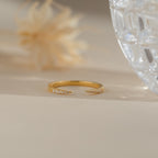 The Jessie Duo Pave Ring, a delicate open ring with sparkling pavé diamonds, is displayed on a beige surface beside a glass and a dried flower.