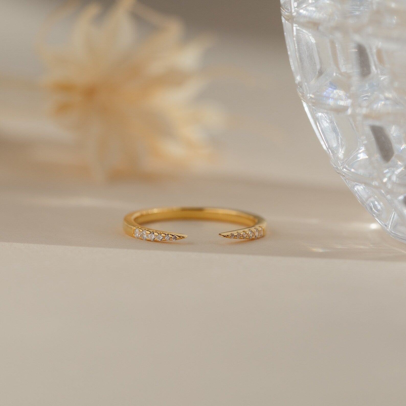 The Jessie Duo Pave Ring, a delicate open ring with sparkling pavé diamonds, is displayed on a beige surface beside a glass and a dried flower.