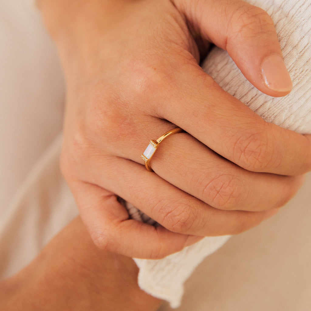 A hand showcases the Kiera Opal Baguette Ring, a dainty design with a gold band and shimmering opal gemstone for an elegant touch.