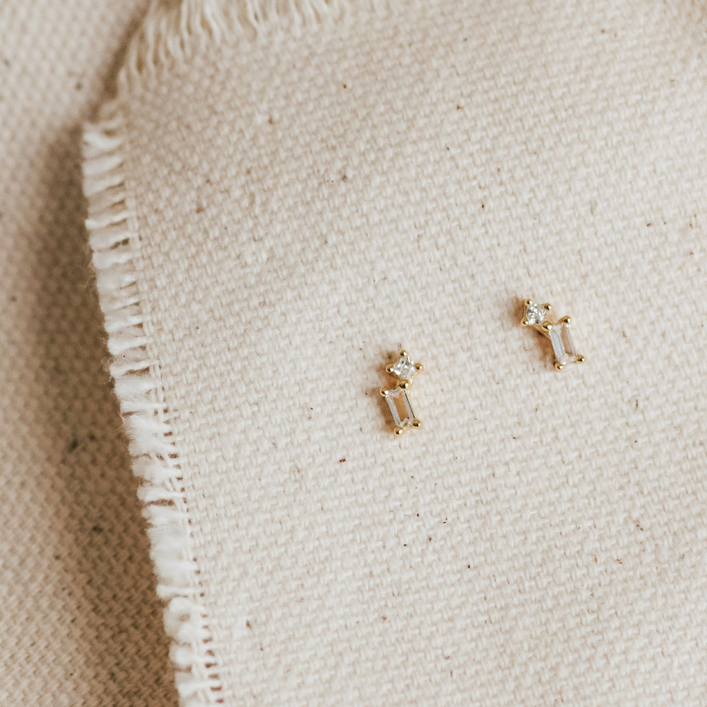 The Blair Baguette Diamond Drop Studs, featuring minimalist clear stones set in gold, rest elegantly on textured cream-colored fabric.