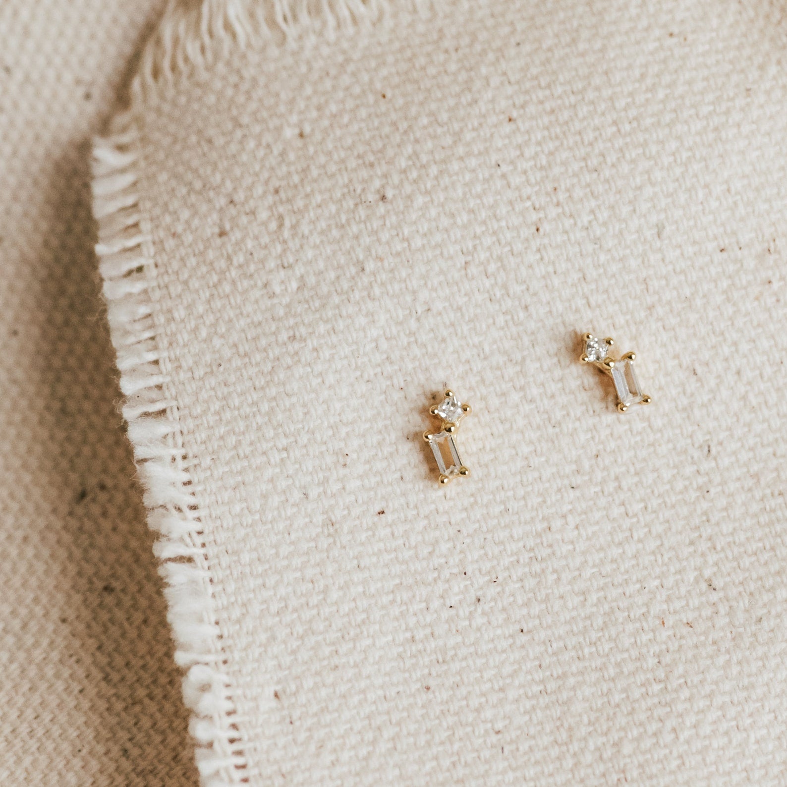 The Blair Baguette Diamond Drop Studs, featuring minimalist clear stones set in gold, rest elegantly on textured cream-colored fabric.