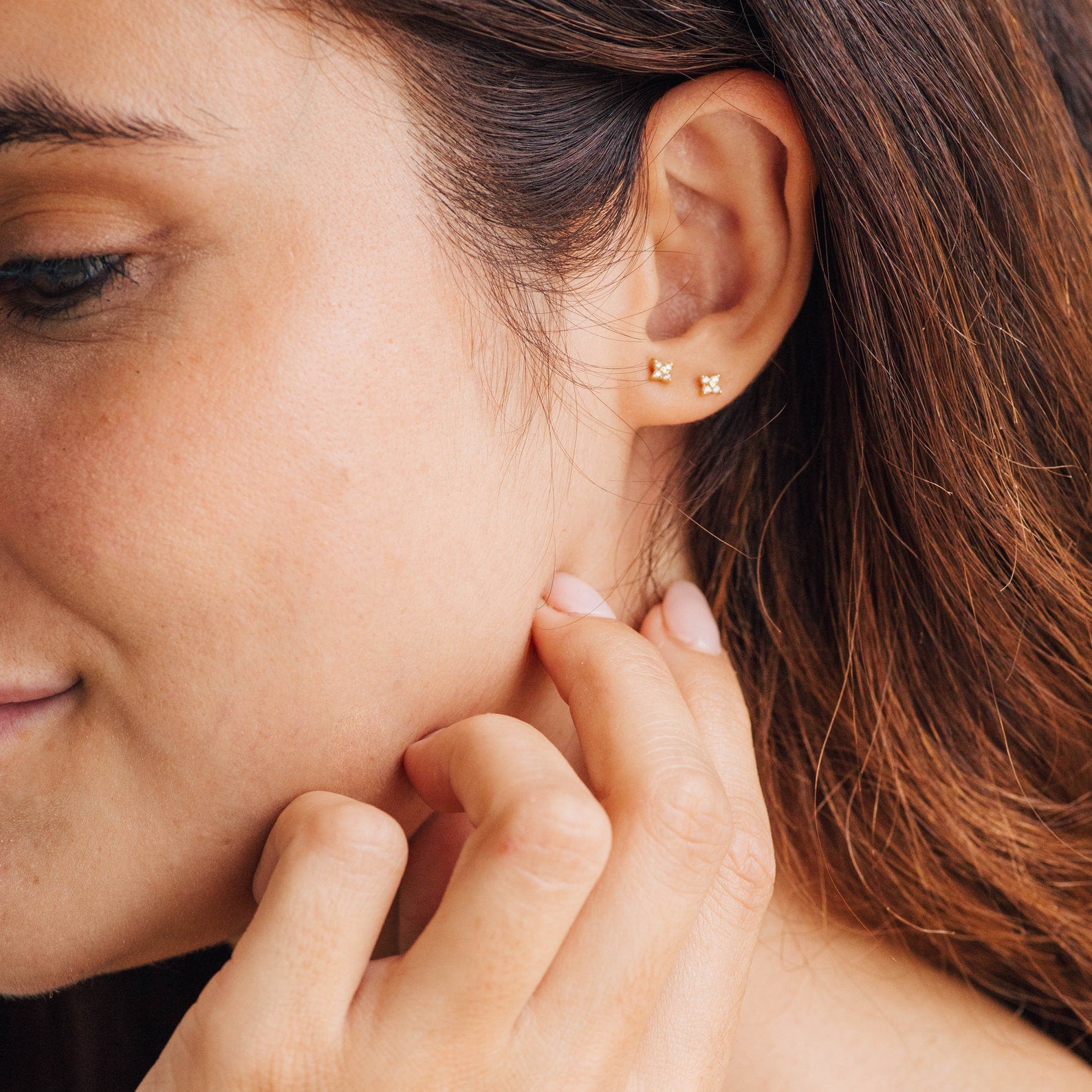 Close-up of a woman smiling and touching her neck while wearing two of our Hera Flower Stud earrings on one earlobe, with her brown hair tucked behind her ear.