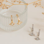 Aleah Opal Evil Eye Huggies—gold and silver huggie hoop earrings with opal gemstone evil eye details, displayed on a glass dish and cream surface. A stunning piece of evil eye jewelry.