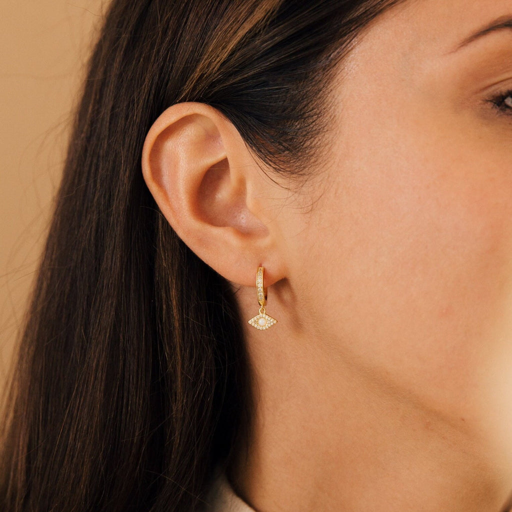 Close-up of a woman's ear wearing Aleah Opal Evil Eye Huggies, featuring a diamond-shaped pendant that blends classic style with modern elegance.