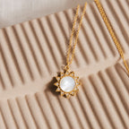 The Elia Sun Necklace features a sun-shaped gold pendant with a white mother of pearl center, displayed on a textured beige surface.