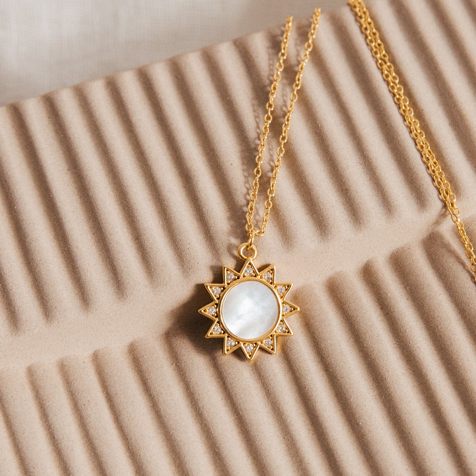 The Elia Sun Necklace features a sun-shaped gold pendant with a white mother of pearl center, displayed on a textured beige surface.
