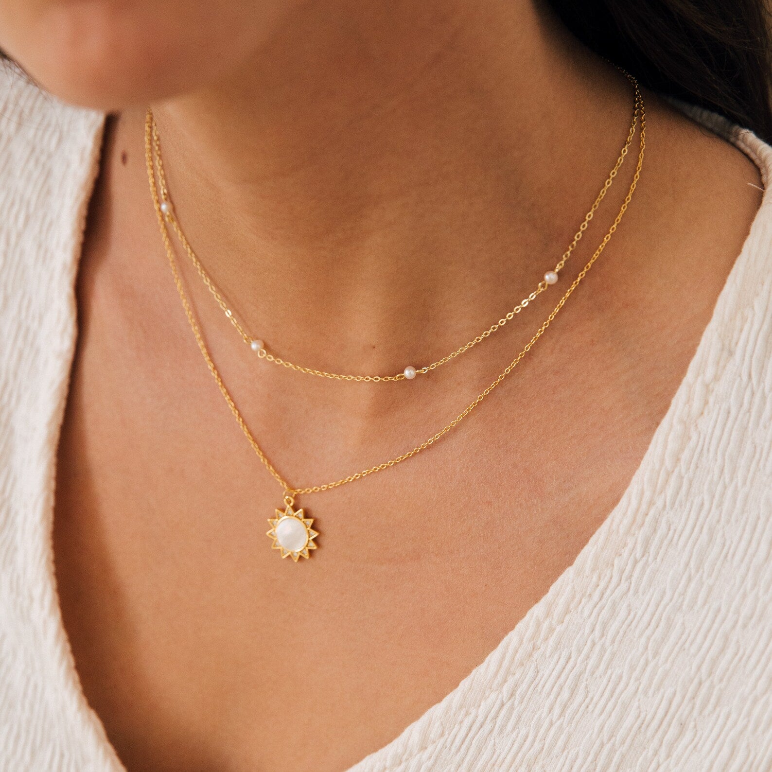 Close-up of a woman wearing two layered gold necklaces, featuring the Dainty Pearl Station Necklace over a textured white top. A perfect gift for mom.