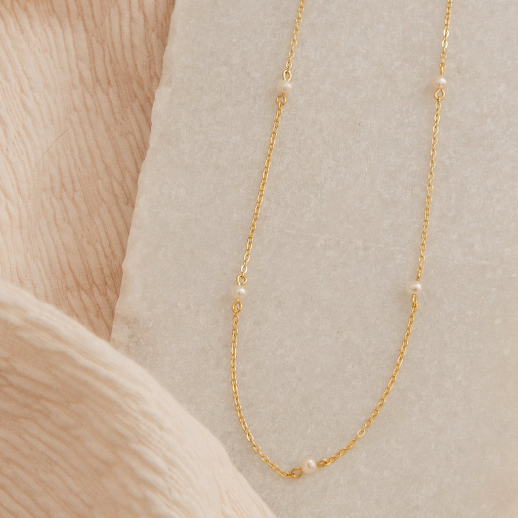 Gold chain necklace with small evenly spaced pearls displayed on a textured cream and beige fabric background.
