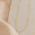 Gold chain necklace with small evenly spaced pearls displayed on a textured cream and beige fabric background.
