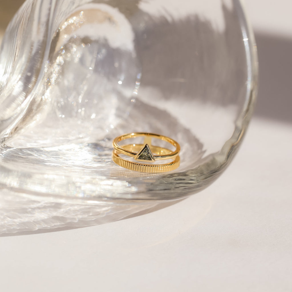 The Jaida Peridot Duo Ring, a gold ring featuring peridot stones, rests under a clear curved glass object on a white surface.