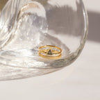 The Jaida Peridot Duo Ring, a gold ring featuring peridot stones, rests under a clear curved glass object on a white surface.