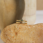 The Jaida Peridot Duo Ring, featuring an elegant Art Deco design with a triangular stone, rests on a textured beige rock against a blurred background.