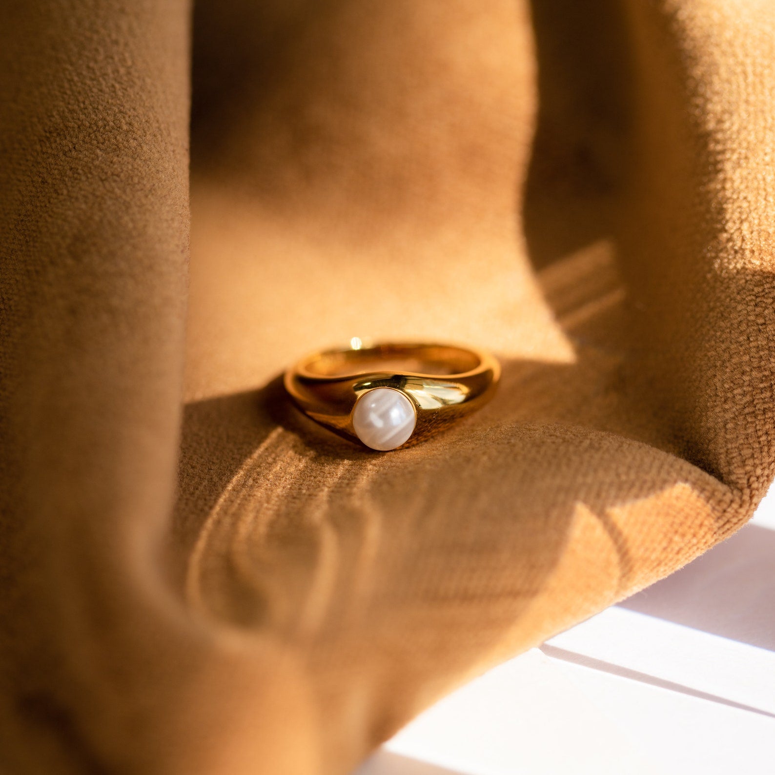 The Penelope Pearl Signet Ring, featuring a single freshwater pearl, rests on soft brown fabric in warm sunlight, capturing the elegance and vintage charm of classic jewelry.