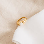 The Penelope Pearl Signet Ring, a piece of gold vintage jewelry with a white freshwater pearl, rests gracefully on a soft cream-colored fabric background.