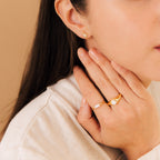 A woman wears gold rings, featuring the Penelope Pearl Signet Ring, and a star-shaped gold earring as her hand gently touches her neck and face.