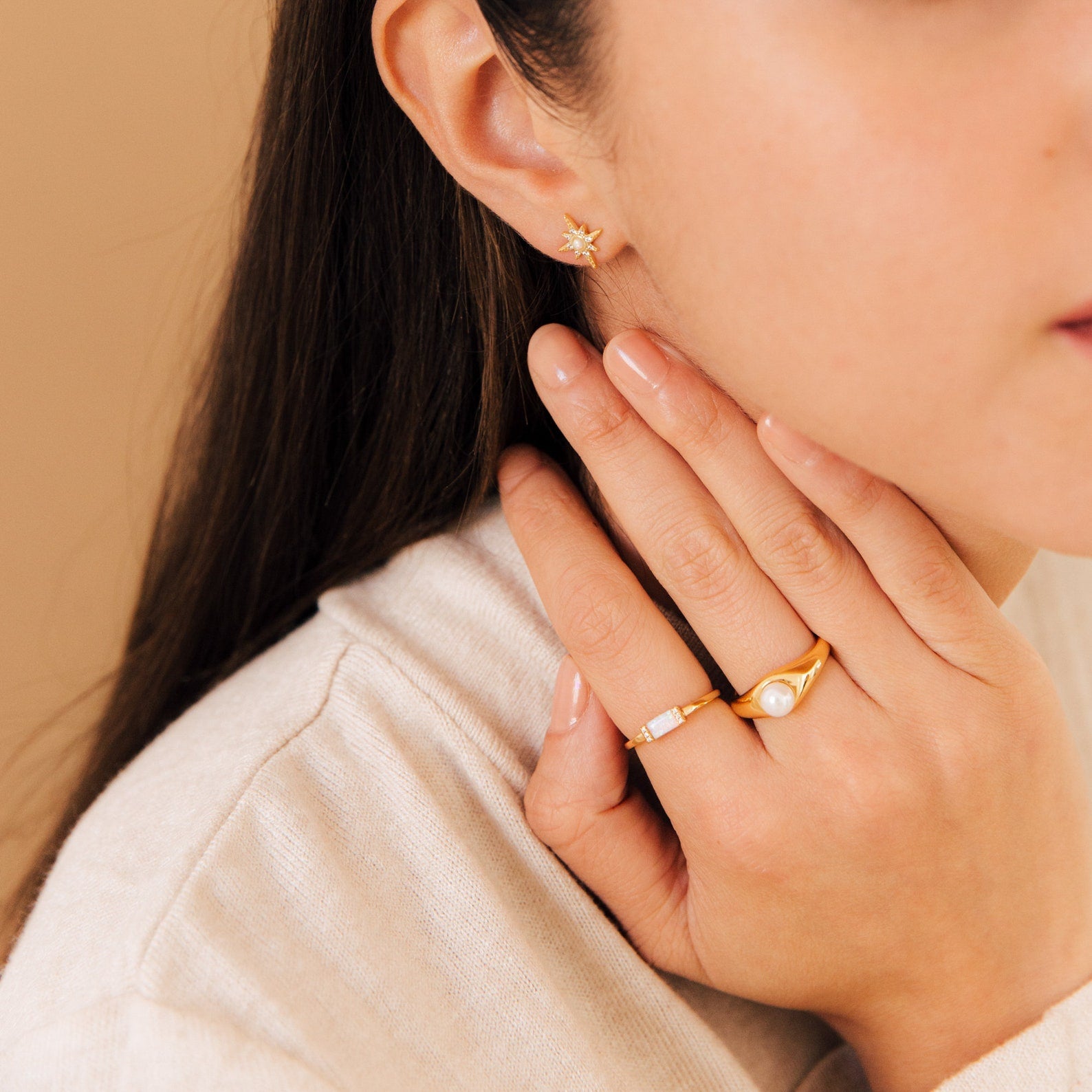 A woman wears gold rings, featuring the Penelope Pearl Signet Ring, and a star-shaped gold earring as her hand gently touches her neck and face.