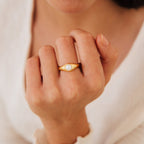 A hand showcases the Penelope Pearl Signet Ring, featuring a central freshwater pearl in a gold setting, set against a soft beige fabric background.