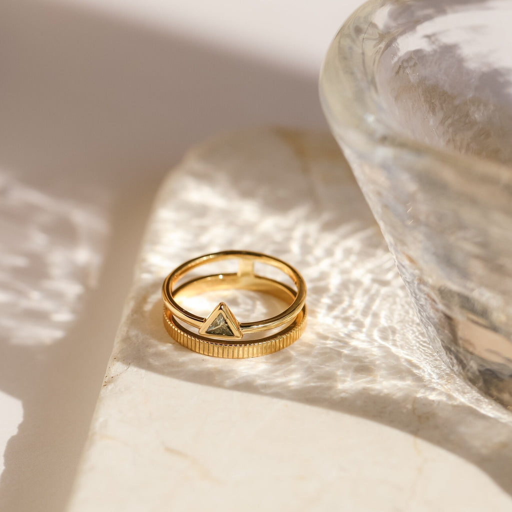 The Jaida Peridot Duo Ring, a gold band featuring two peridot stones, rests on a marble surface in soft light near a glass bowl.