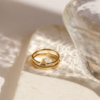 The Jaida Peridot Duo Ring, a gold band featuring two peridot stones, rests on a marble surface in soft light near a glass bowl.