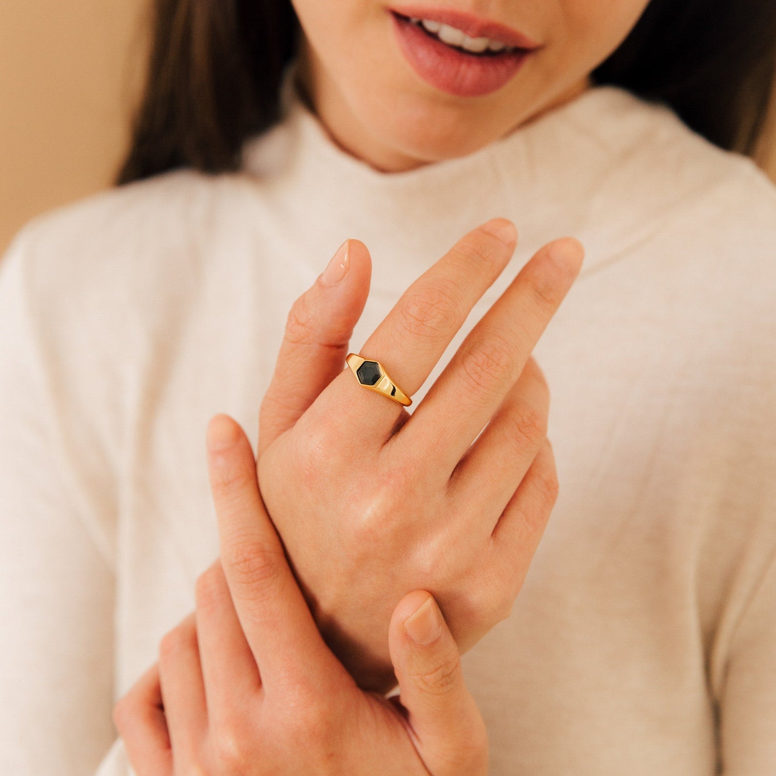 A woman in a cream top gently touches her hand, showcasing the Ember Signet Ring—a bold gold ring with a striking black stone that adds an elegant touch to her style.