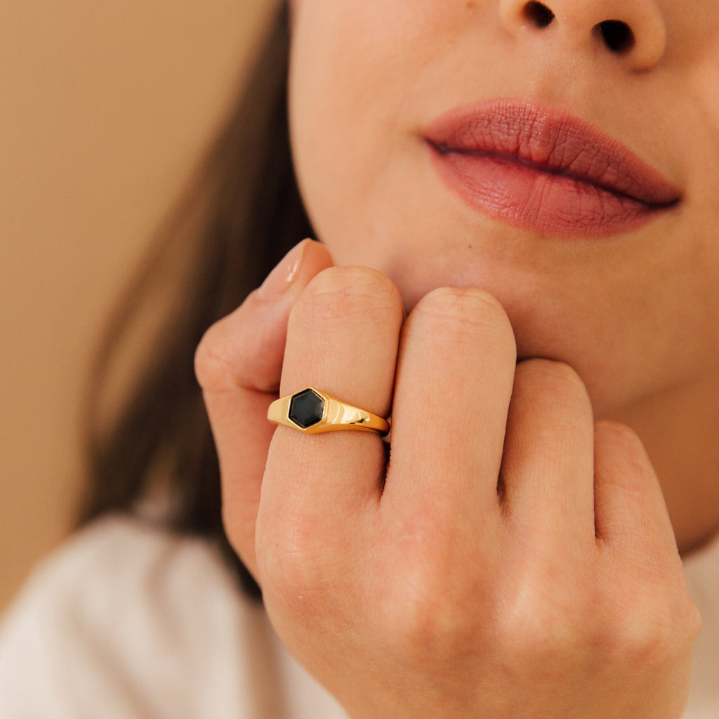 A woman with natural makeup wears the Ember Signet Ring, a chunky gold and black signet design, as she rests her hand on her chin.