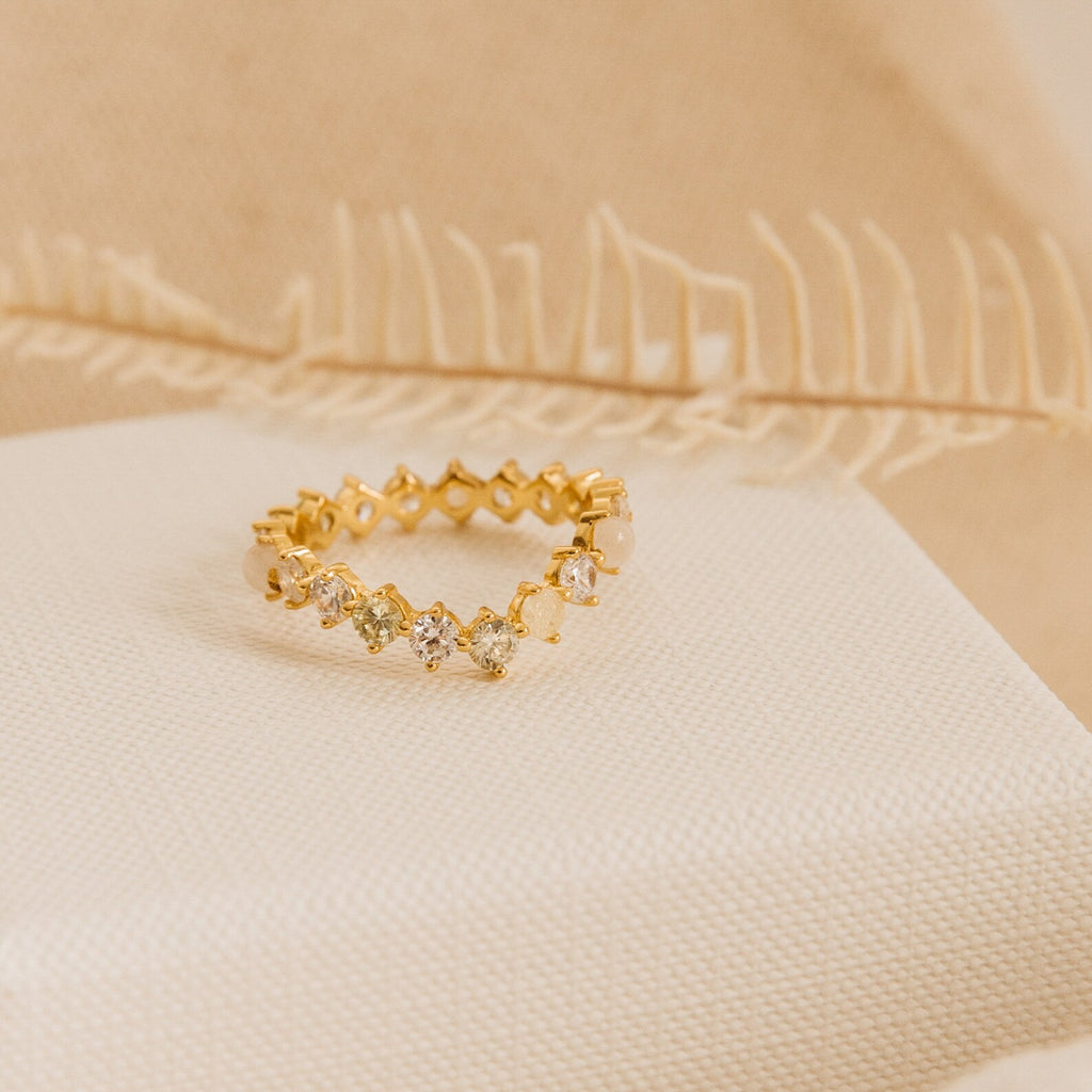 The Mosaic Wave Eternity Ring, a gold band with small diamonds, is displayed on cream fabric. This unique ring sparkles delicately like a moonstone diamond eternity band against a soft beige background.