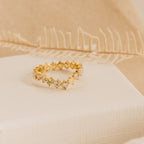 The Mosaic Wave Eternity Ring, a gold band with small diamonds, is displayed on cream fabric. This unique ring sparkles delicately like a moonstone diamond eternity band against a soft beige background.