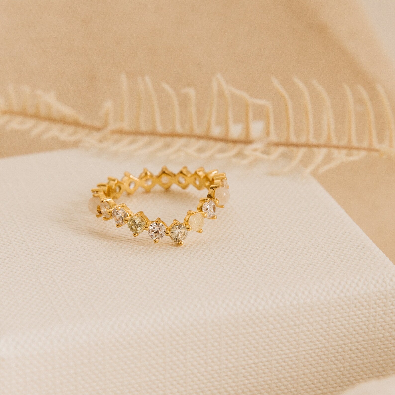 The Mosaic Wave Eternity Ring, a gold band with small diamonds, is displayed on cream fabric. This unique ring sparkles delicately like a moonstone diamond eternity band against a soft beige background.