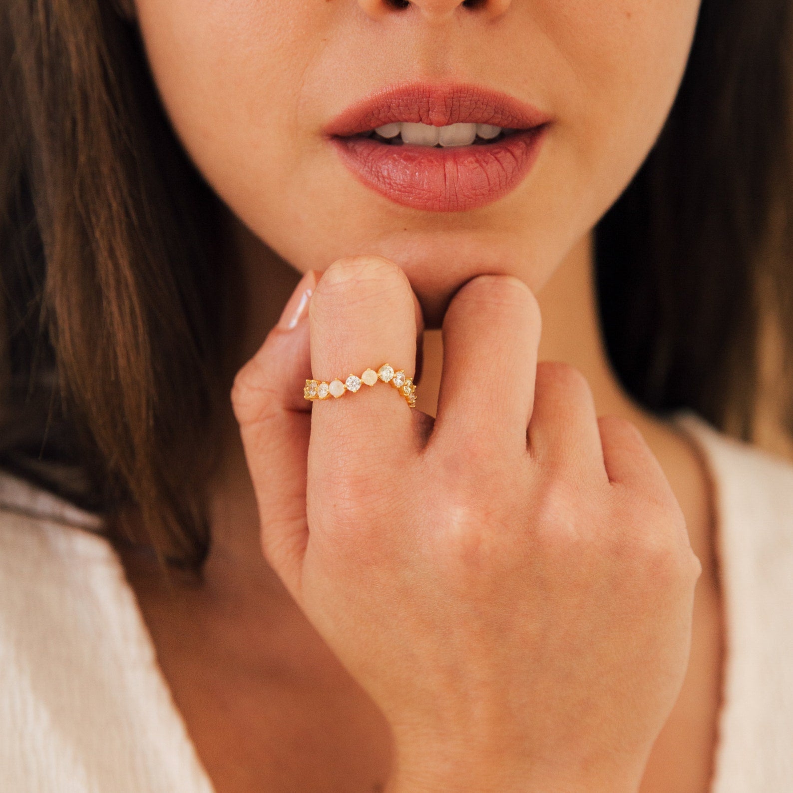 A woman holds her hand to her face, showcasing the Mosaic Wave Eternity Ring—a gold band decorated with sparkling diamond-like stones on her finger.