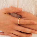Close-up of hands wearing the Mosaic Wave Eternity Ring, a delicate gold band adorned with small white stones, shown against a cream fabric background.