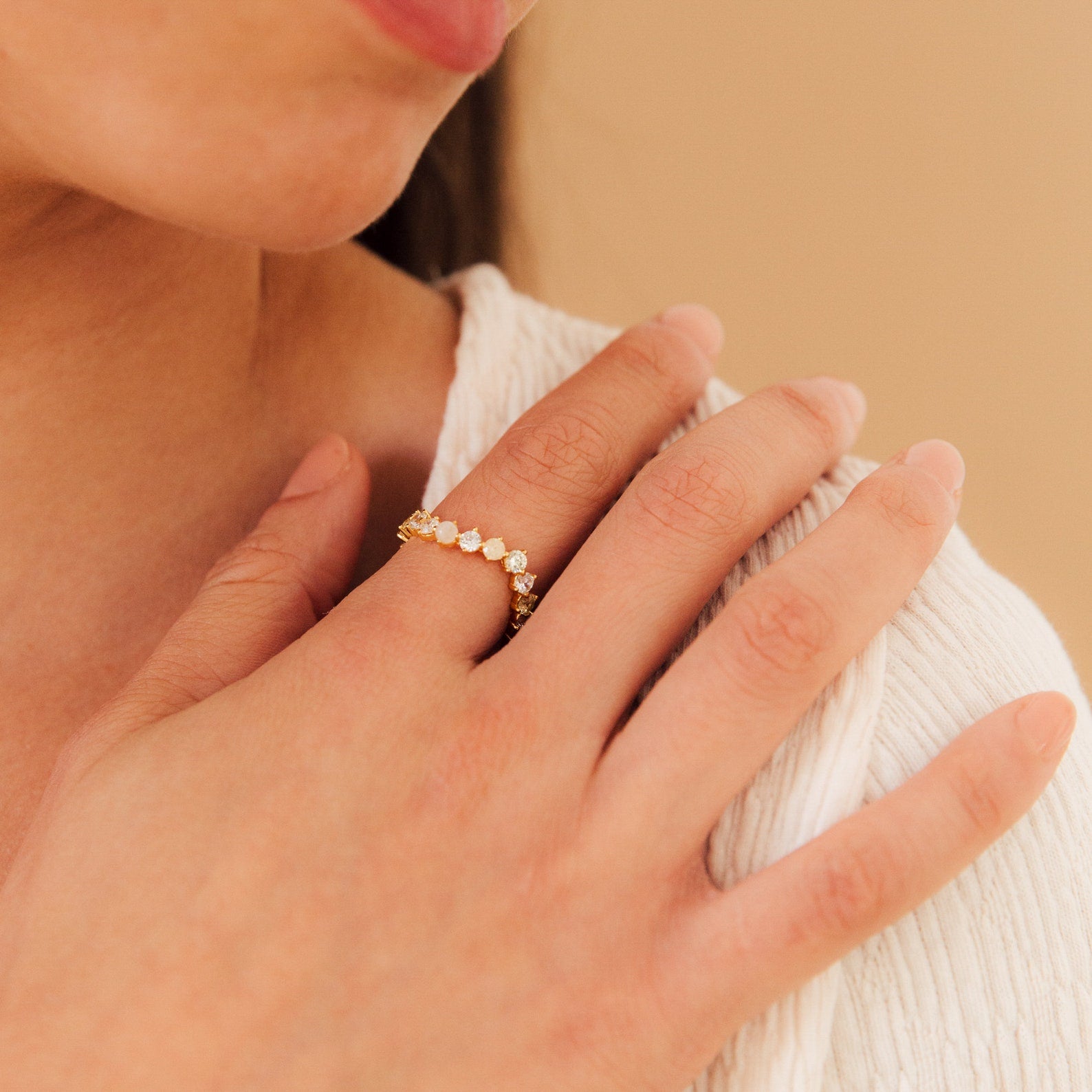 A woman’s hand with the Mosaic Wave Eternity Ring—a gold band set with moonstone and sparkling white stones—rests near her collarbone.