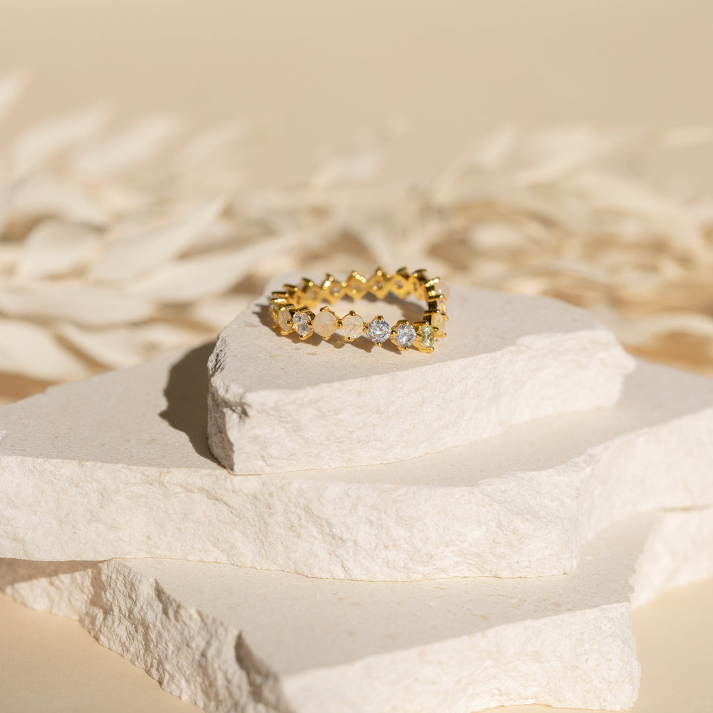 The Mosaic Wave Eternity Ring, featuring alternating round gemstones, shines atop textured white stone slabs against a beige background.