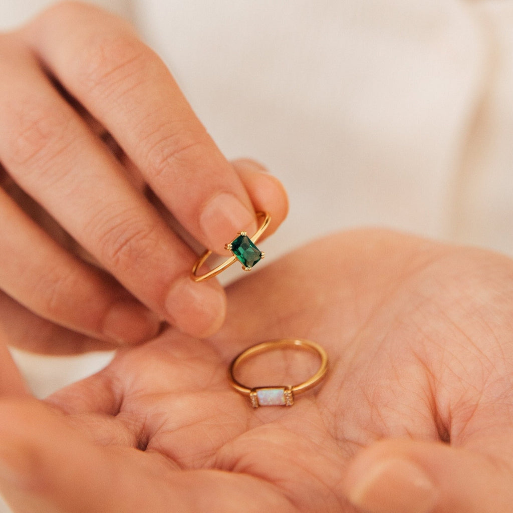 A hand displays the Harley Emerald Ring, featuring a vivid green gemstone, while another art deco ring rests on the other palm.
