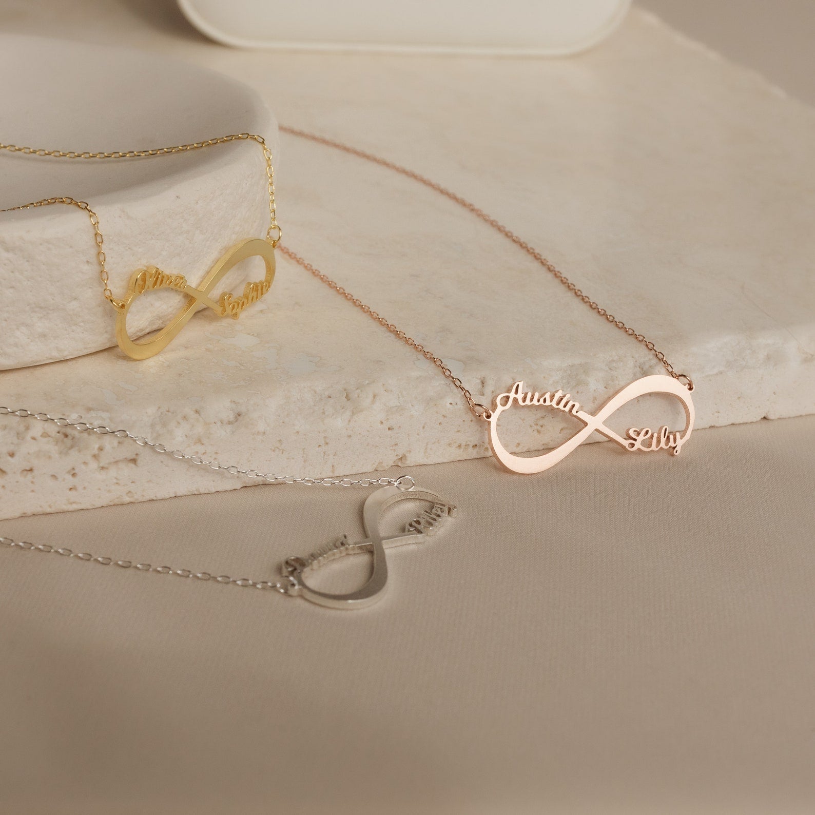 The Infinity Name Necklace, shown in gold, rose gold, and silver on a neutral stone, makes a perfect personalized necklace and thoughtful anniversary gift.