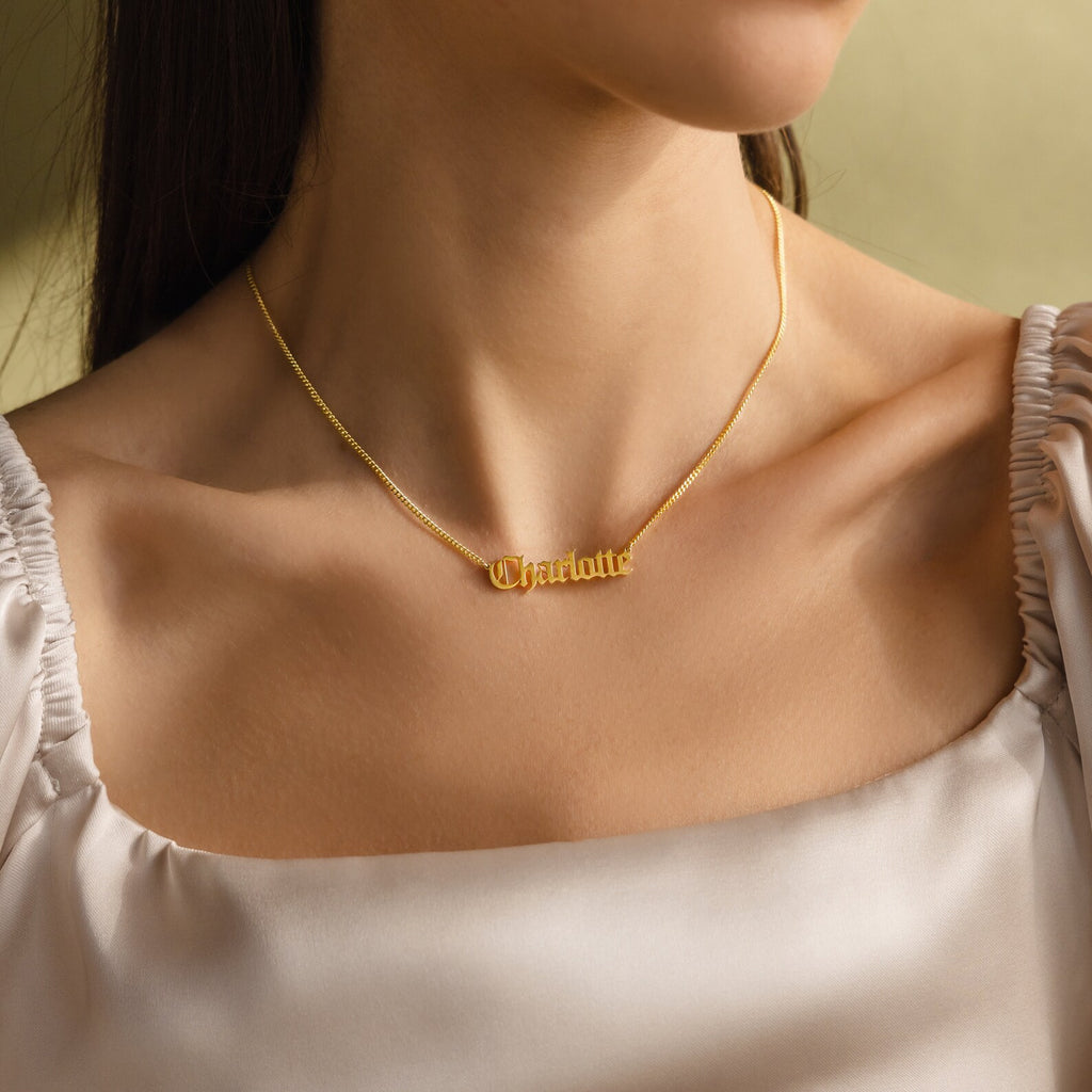 A woman in a cream-colored top wears the Old English Name Necklace in Curb Chain, featuring "Charlotte" in gold script.