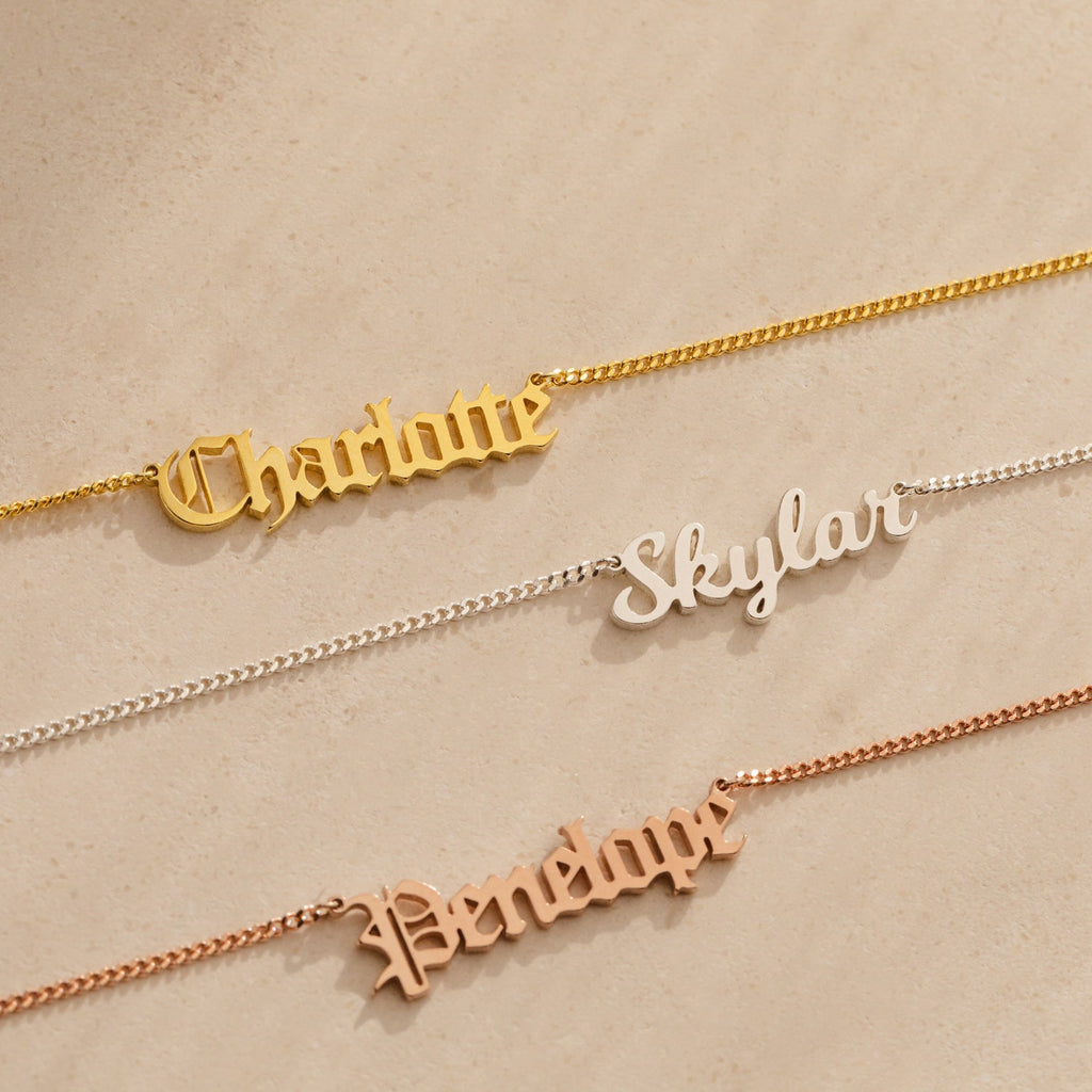 Three Old English Name Necklaces in curb chains lay on a beige surface, personalized with "Charlotte," "Skylar," and "Penelope" in different metals, each featuring a custom charm for a modern look.