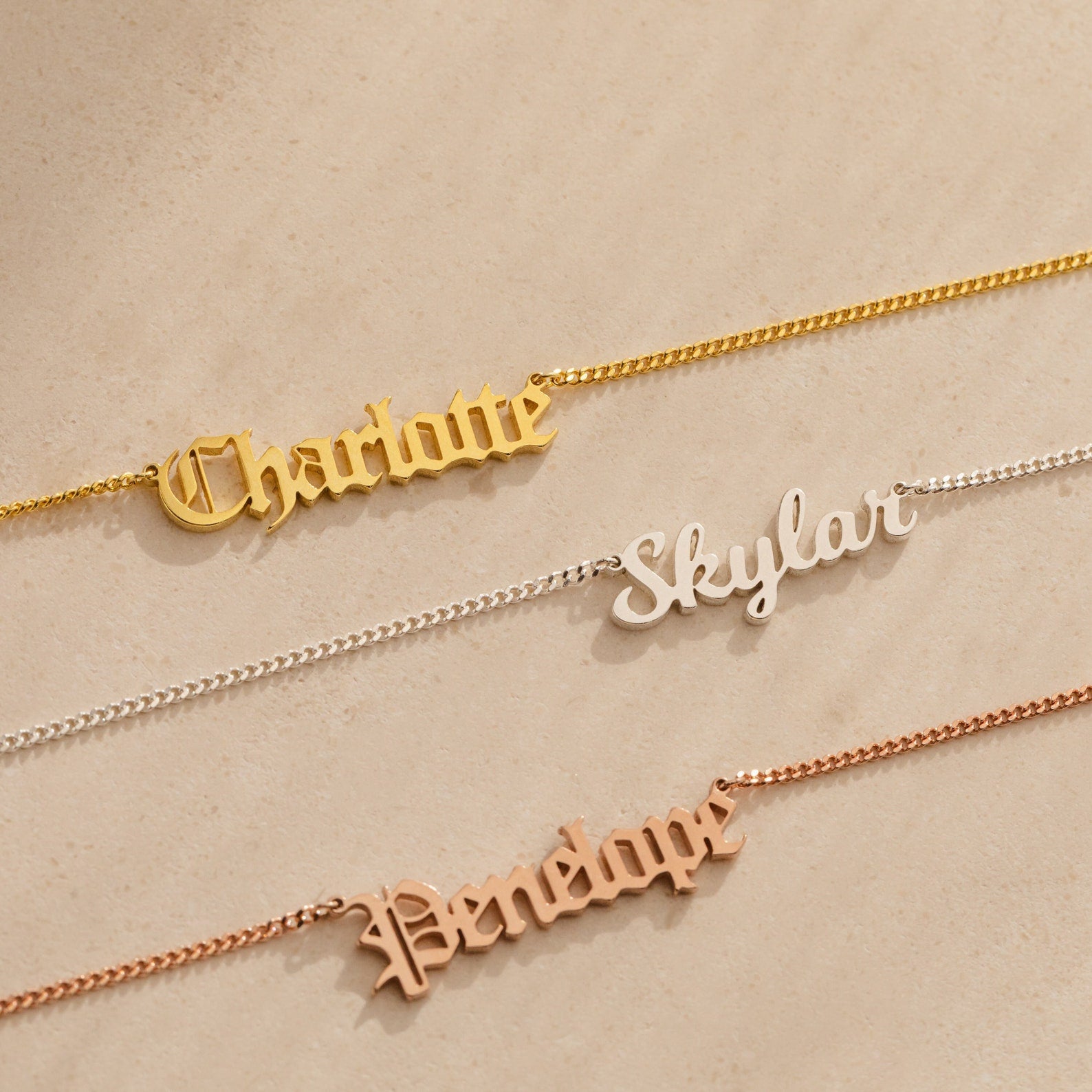 Three Old English Name Necklaces in curb chains lay on a beige surface, personalized with "Charlotte," "Skylar," and "Penelope" in different metals, each featuring a custom charm for a modern look.