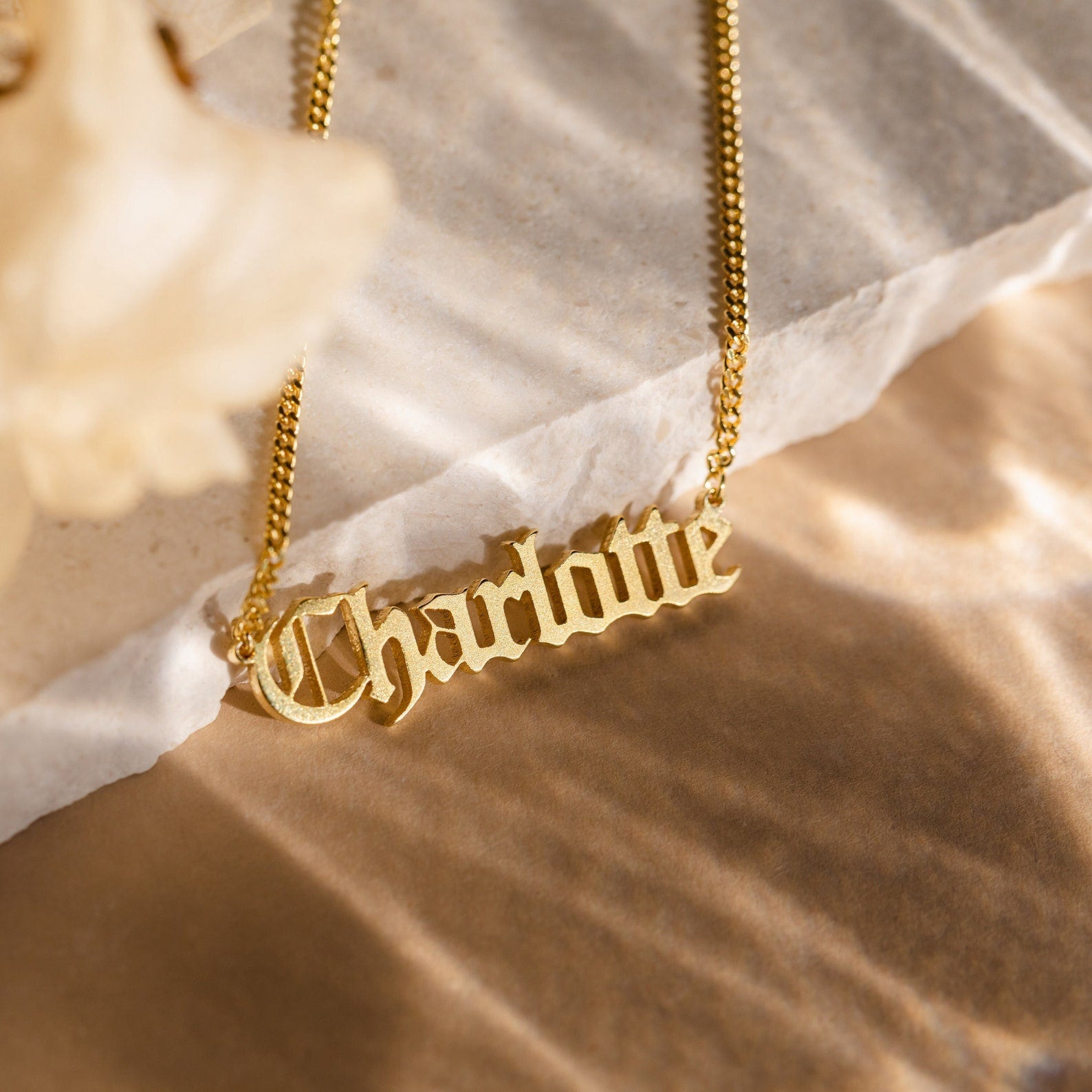 The Old English Name Necklace in Curb Chain features a gothic nameplate reading Charlotte, displayed on a marble surface.