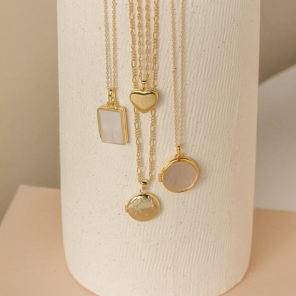 Four Initial Heart Locket Necklaces, displayed vertically on a textured white cylinder—perfect as personalized jewelry.