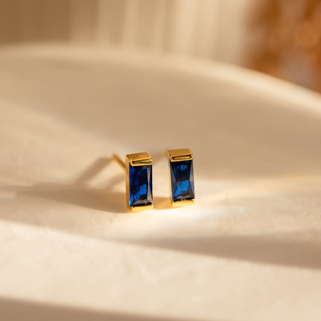 Baguette Birthstone Studs: Rectangular gold earrings with deep blue gems on a smooth, light surface—ideal as elegant birthstone studs or personalized gifts.