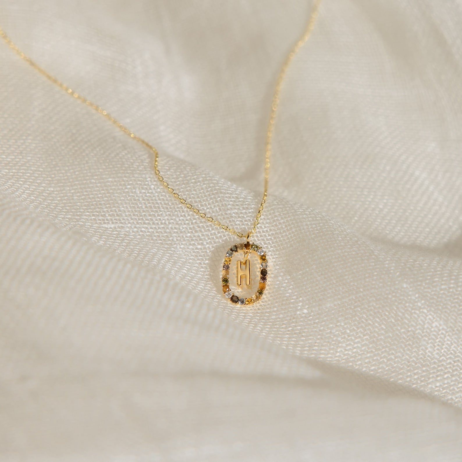 Rainbow Initial Necklace featuring a round pendant engraved with the letter M, displayed on soft white fabric—a timeless personalized piece perfect for adding a special touch to any style.