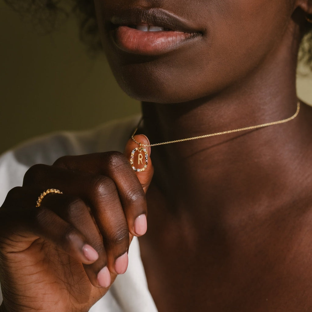 A woman holds a Rainbow Initial Necklace with a gold “R” pendant close to her lips.