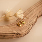 Reagan Duo Huggies, gold geometric double hoop dangle earrings, rest on a textured wooden branch with dried flowers in the background, creating a chic and elegant look.