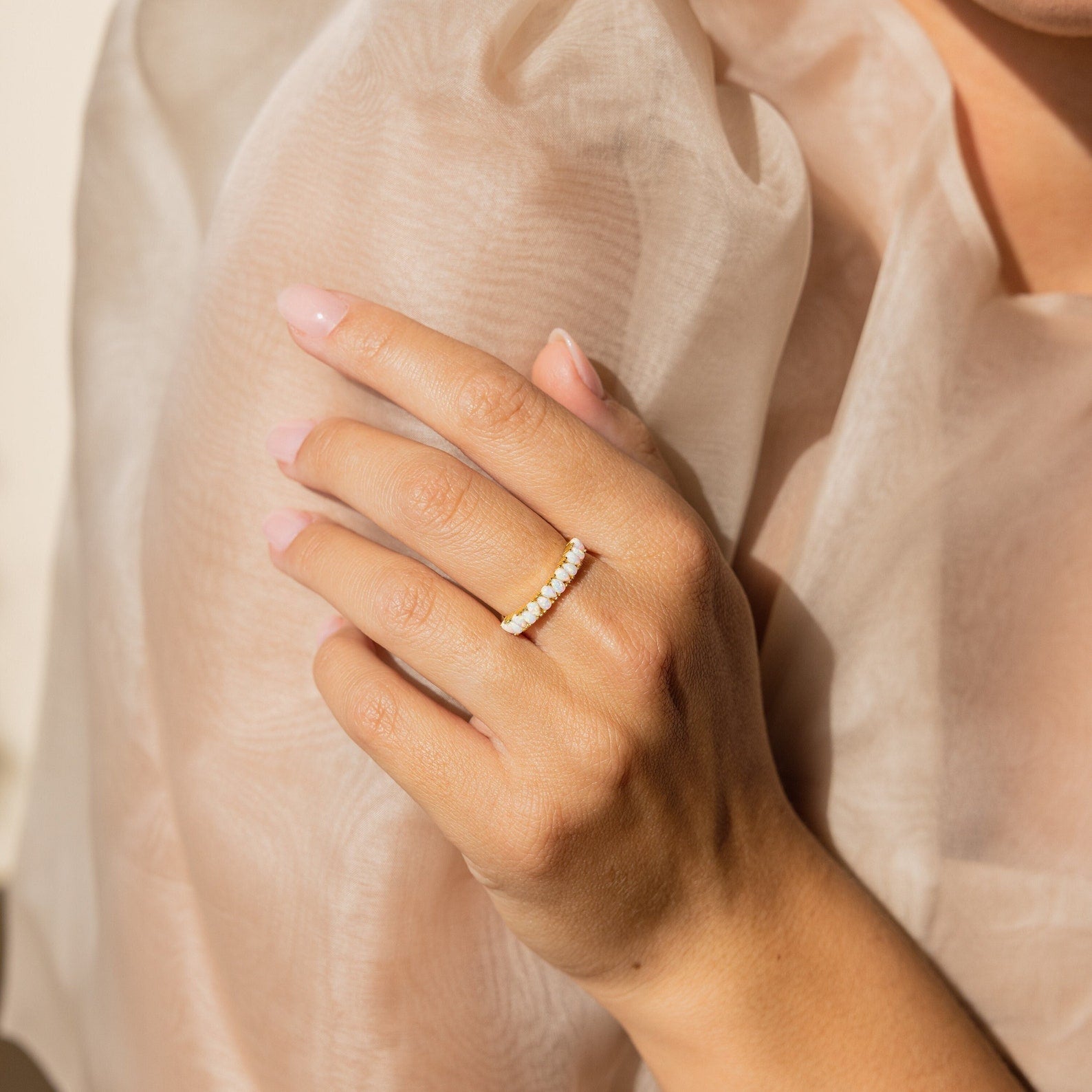 A hand with light pink nails wears the vintage-inspired Opal Drop Eternity Ring and small pearls, resting on sheer beige fabric.