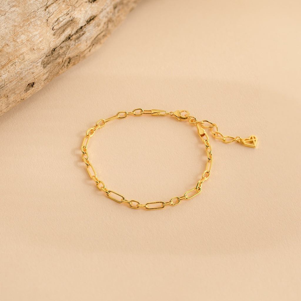 The Roxy Paperclip Chain Bracelet features delicate elongated gold links with a small heart charm, ideal for minimal jewelry lovers and beautifully displayed on a beige background.