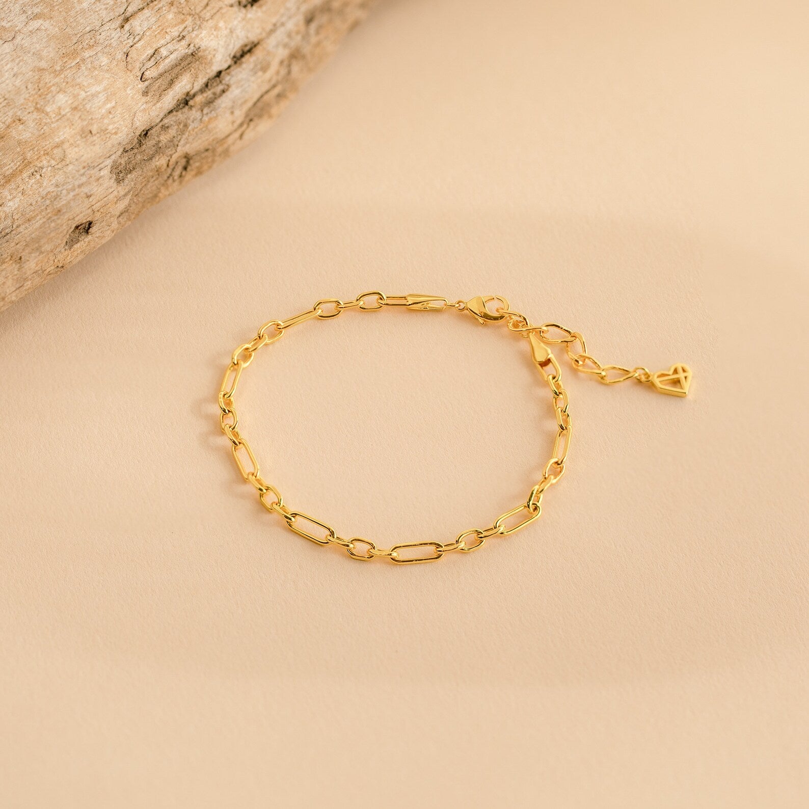 The Roxy Paperclip Chain Bracelet features delicate elongated gold links with a small heart charm, ideal for minimal jewelry lovers and beautifully displayed on a beige background.