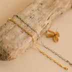 The Roxy Paperclip Chain Bracelet, shown with gold and silver chain necklaces on driftwood, is paired with a dried brown leaf for a minimal jewelry look.