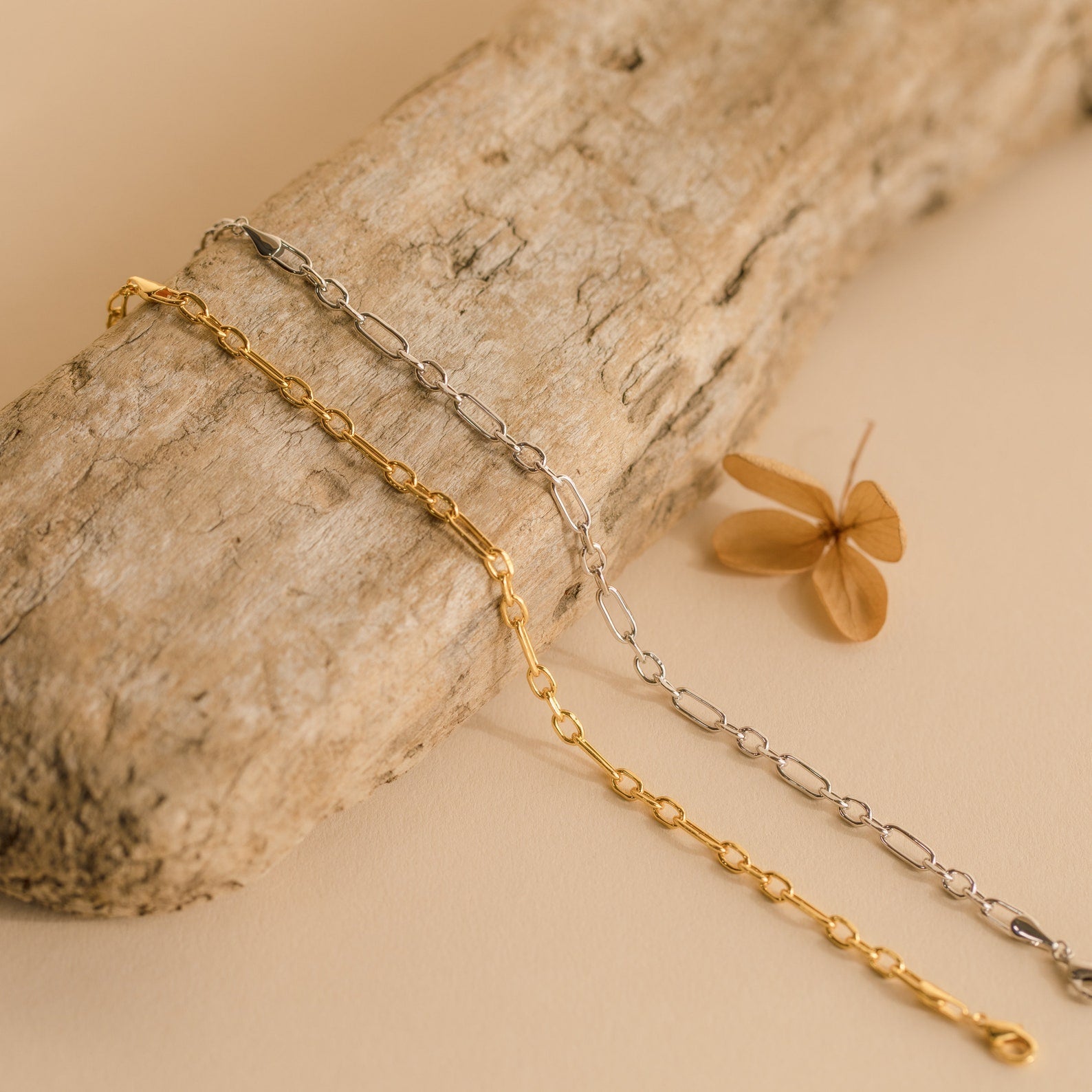 The Roxy Paperclip Chain Bracelet, shown with gold and silver chain necklaces on driftwood, is paired with a dried brown leaf for a minimal jewelry look.